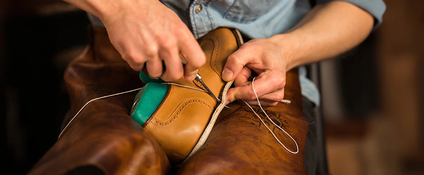 A close-up of a cobbler's hands sewing the sole of a tan leather shoe onto a brown wooden leg form, with a green shoe holder visible.